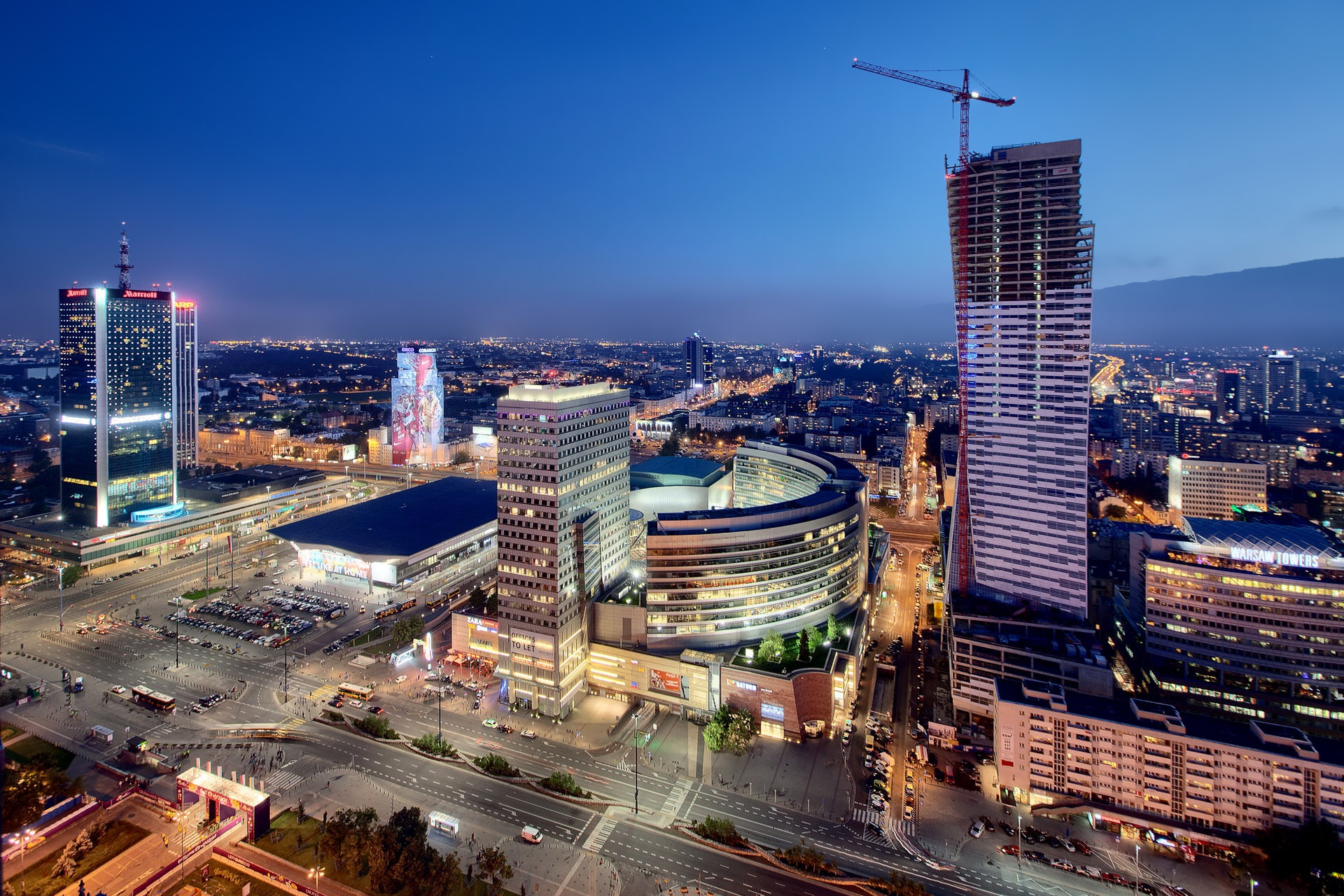 Panorama Warszawy nocą z widokiem na drapacze chmur, w tym Marriott, Złota 44 w budowie z żurawiem, oraz oświetlone ulice i budynki w oddali.