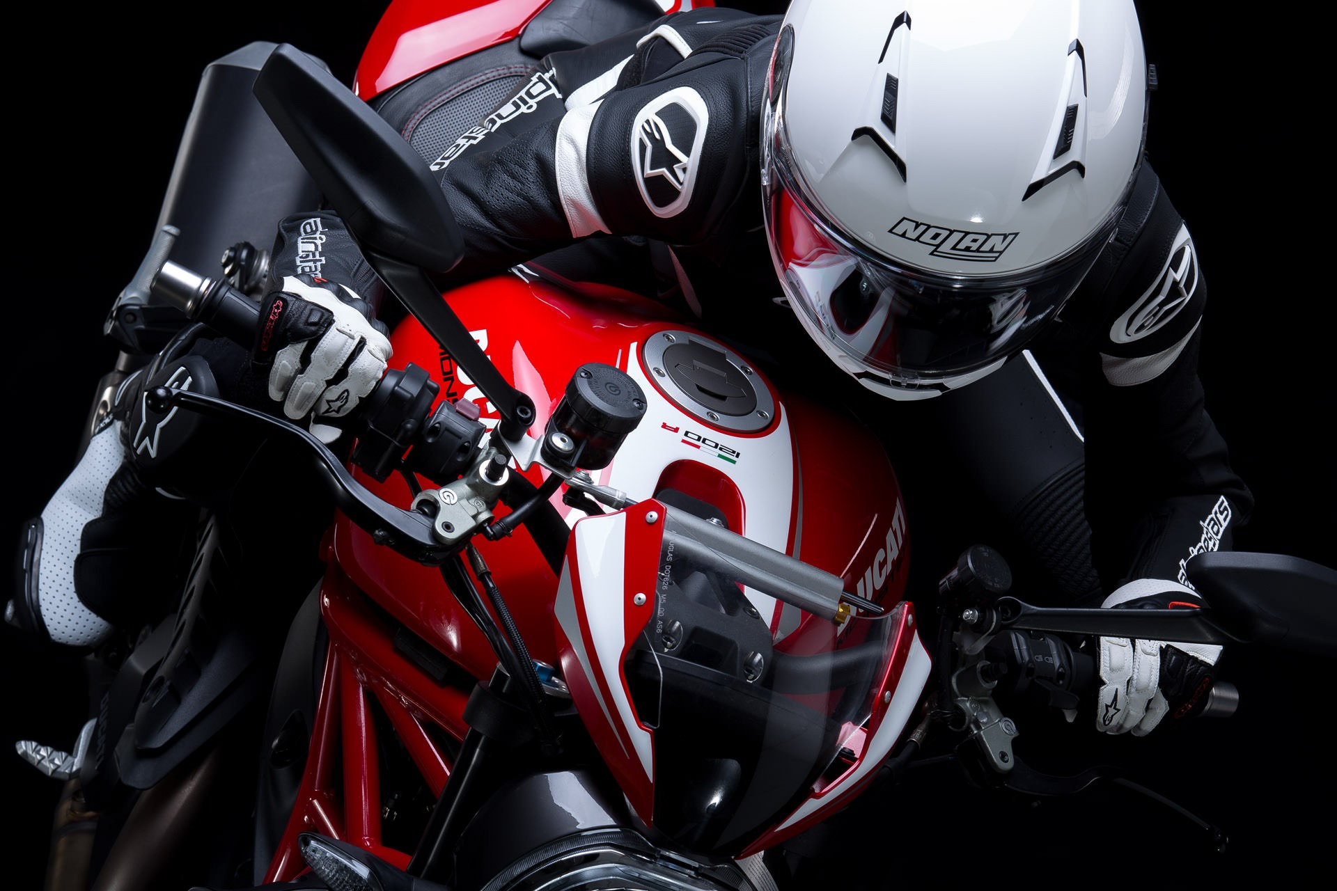 Zbliżenie na motocykl Ducati 1200 R z kierowcą w skórzanym kombinezonie i białym kasku Nolan, ujęcie z góry na czarnym tle.
