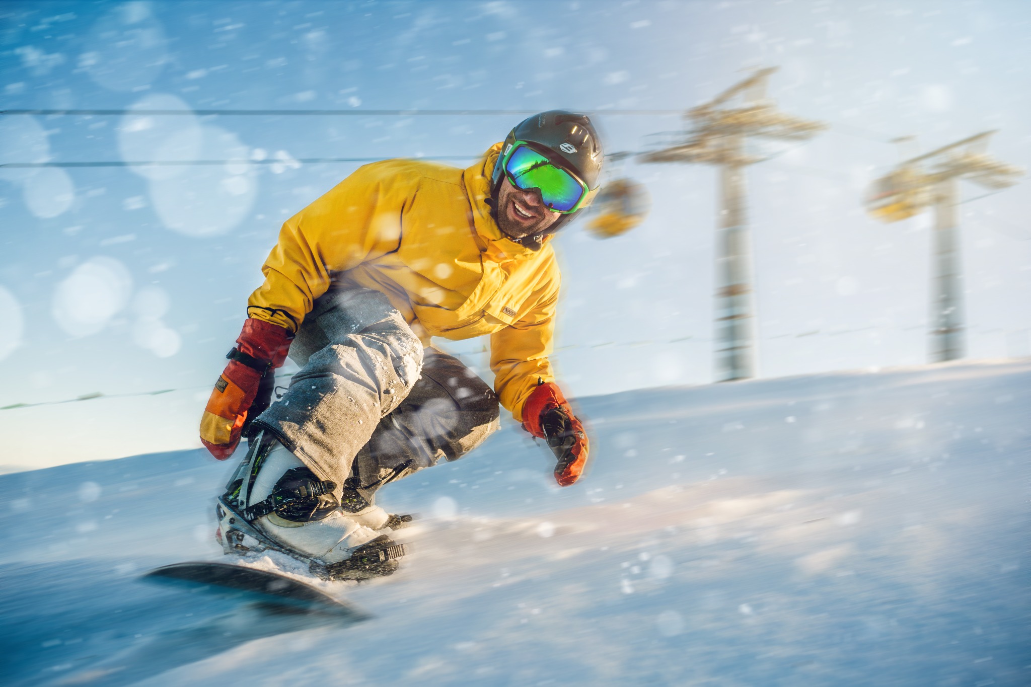 Uśmiechnięty snowboardzista w żółtej kurtce i zielonych goglach zjeżdża po ośnieżonym stoku, z wyciągiem narciarskim w tle.