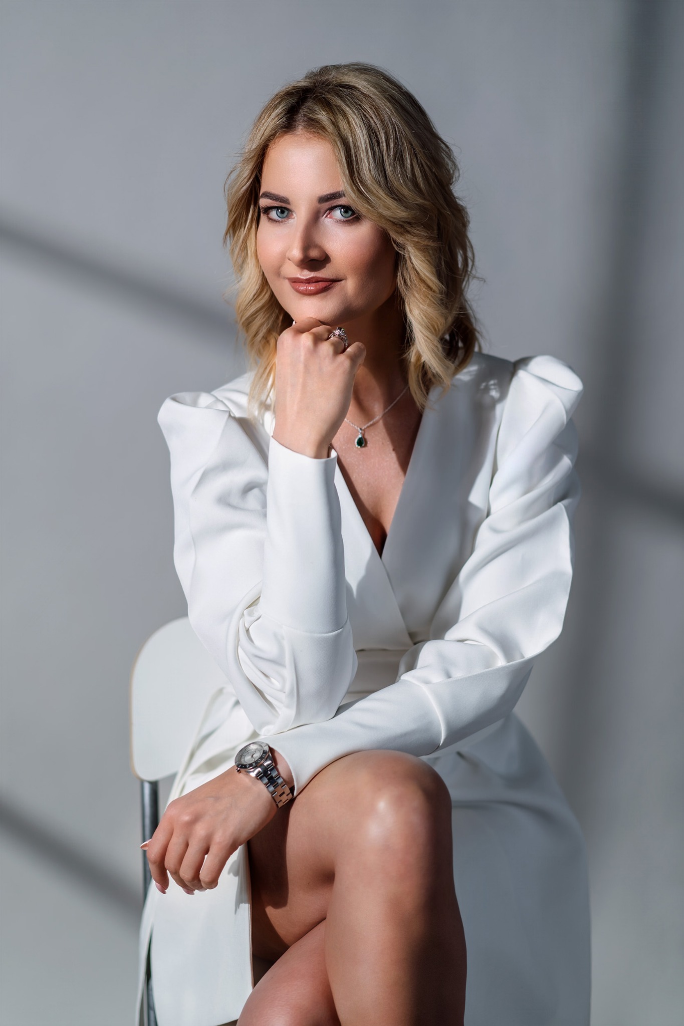 Portret kobiety w białej marynarce siedzącej na krześle, z dłonią opartą o podbródek, z widocznym srebrnym zegarkiem i pierścionkiem z zielonym oczkiem, na jasnoszarym tle z cieniem.