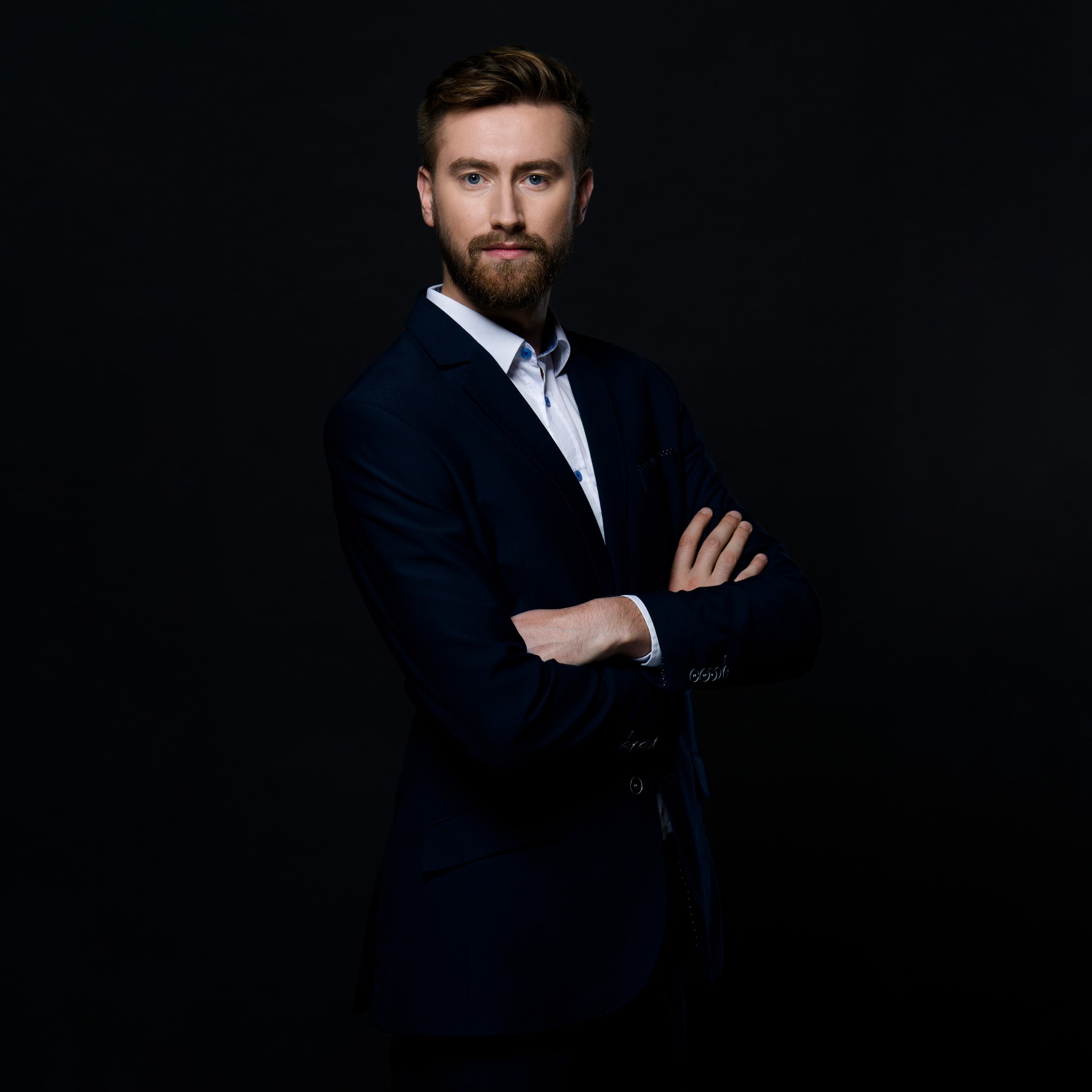 Portret mężczyzny z brodą w granatowym garniturze i białej koszuli, z założonymi rękami na ciemnym tle.