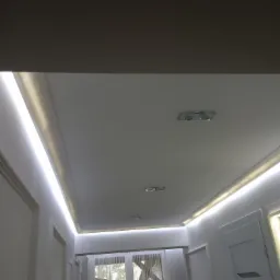 Białe wnętrze z podwieszanym sufitem i oświetleniem LED, widok na okno z firankami.