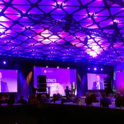 Konferencja firmowa z oświetleniem w odcieniach fioletu, prelegent na scenie i publiczność siedząca przy stołach.