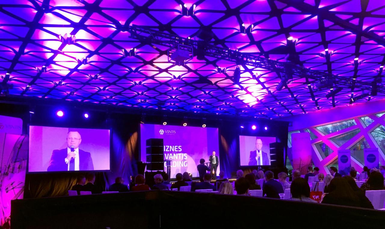 Konferencja firmowa z oświetleniem w odcieniach fioletu, prelegent na scenie i publiczność siedząca przy stołach.