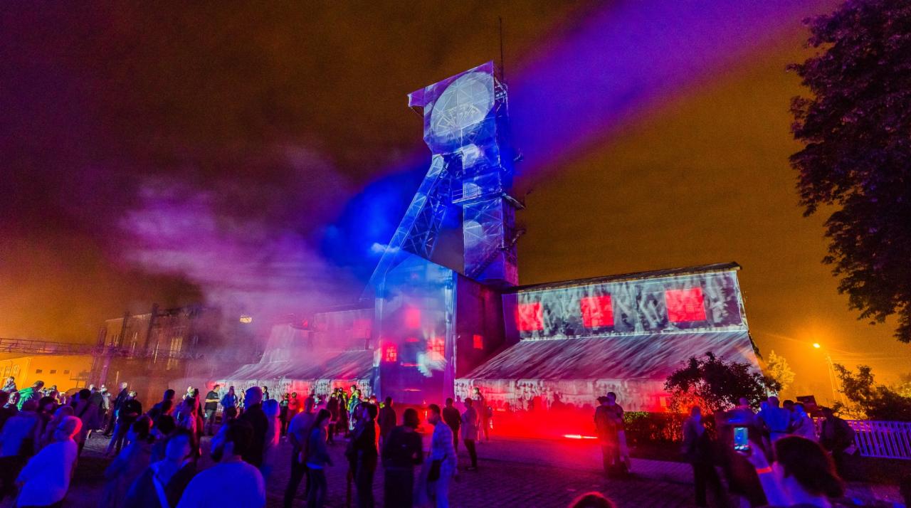Nocny pokaz iluminacji na budynku kopalni, tłum ludzi obserwuje efekty świetlne w kolorach niebieskim, fioletowym i czerwonym.