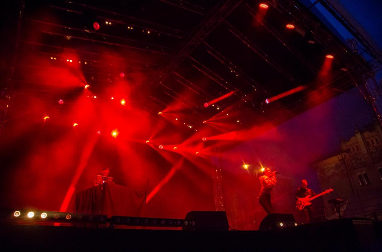 Scena oświetlona czerwonymi światłami z występującym wokalistą, gitarzystą i DJ-em za konsoletą.