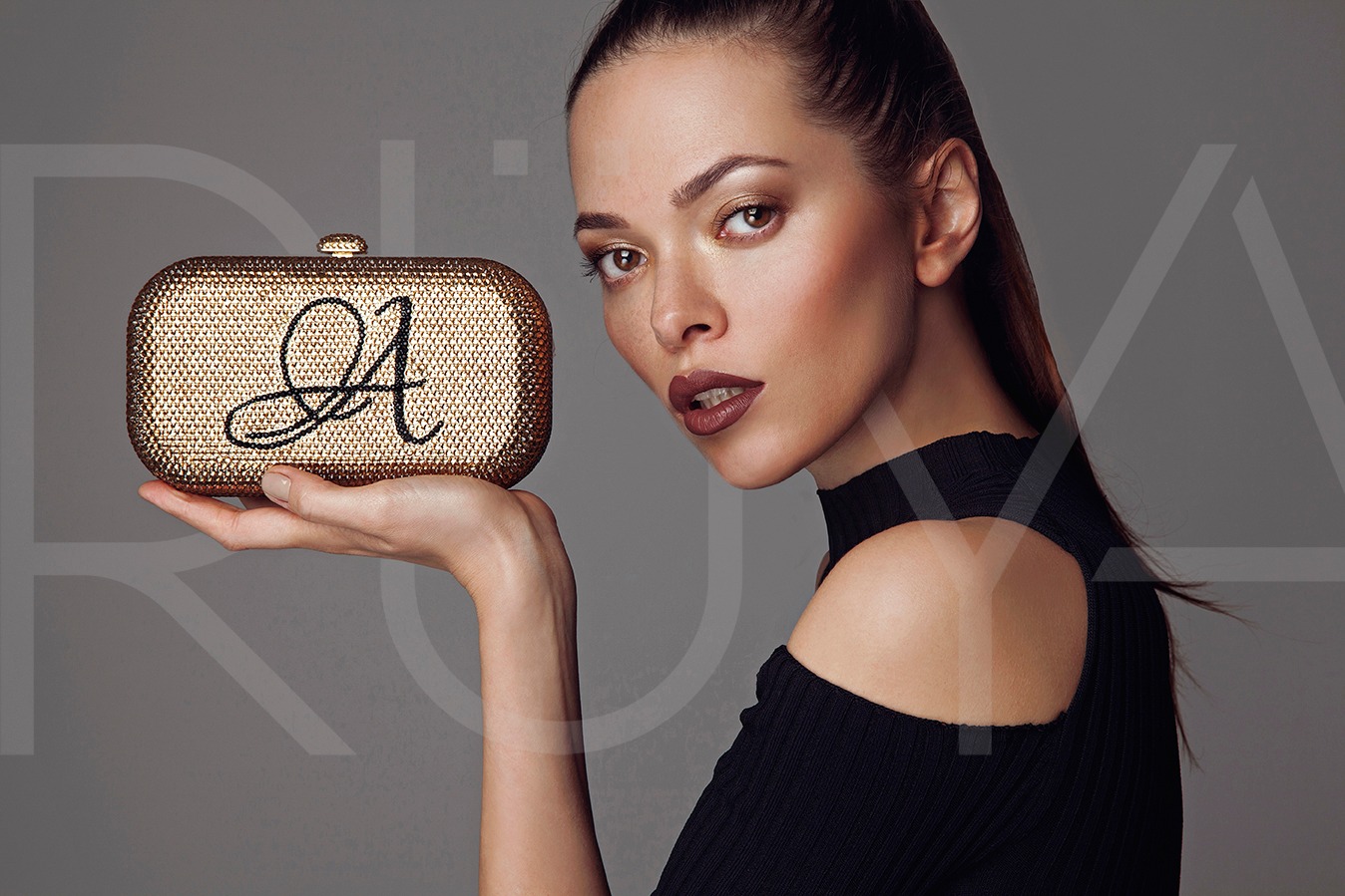 Portret kobiety z błyszczącą, złotą kopertówką z czarnym monogramem 'A', trzymaną na otwartej dłoni, na szarym tle.