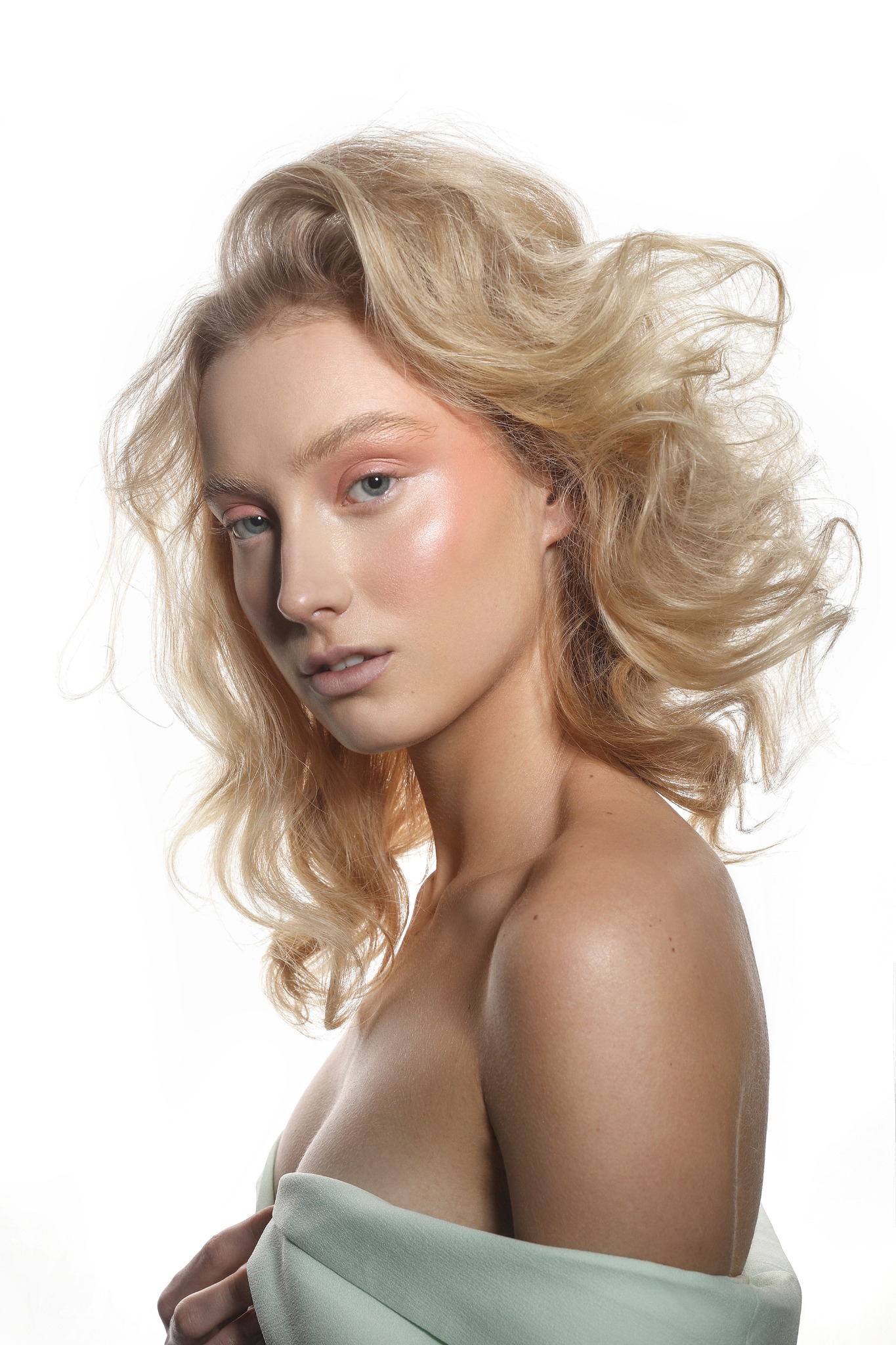 Portret kobiety z blond włosami w artystycznym nieładzie, delikatny makijaż z różowym akcentem na powiekach i policzkach, jasna karnacja, ujęcie z profilu na białym tle.