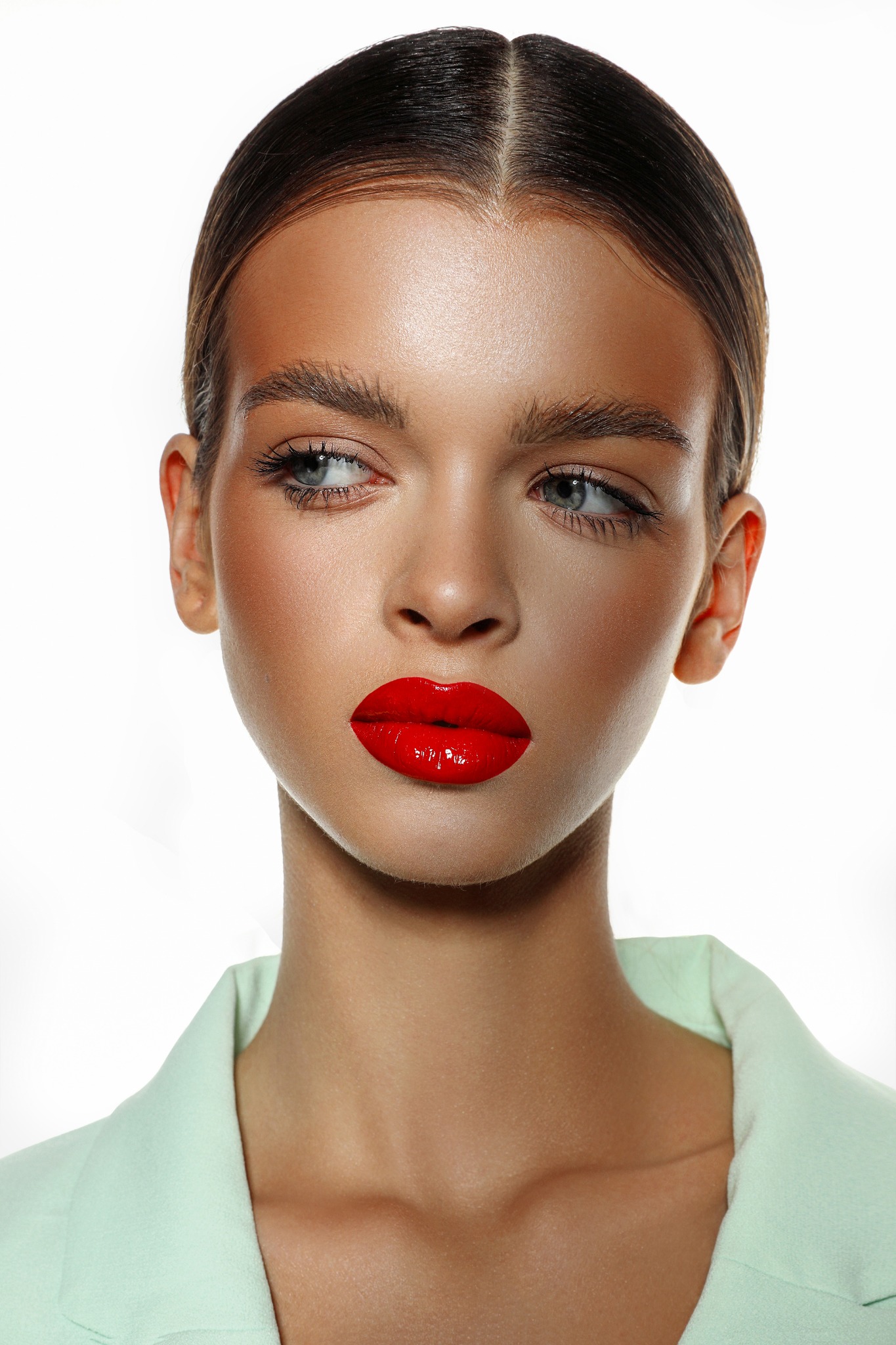 Portret kobiety z mocnym makijażem, błyszczące czerwone usta, subtelny cień do powiek i pastelowy żakiet na białym tle.