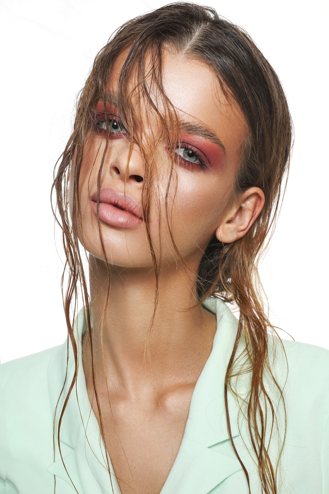 Portret modelki z mokrymi włosami zasłaniającymi częściowo twarz, intensywny makijaż oczu w odcieniach różu, ubrana w jasnozielony żakiet, na białym tle.