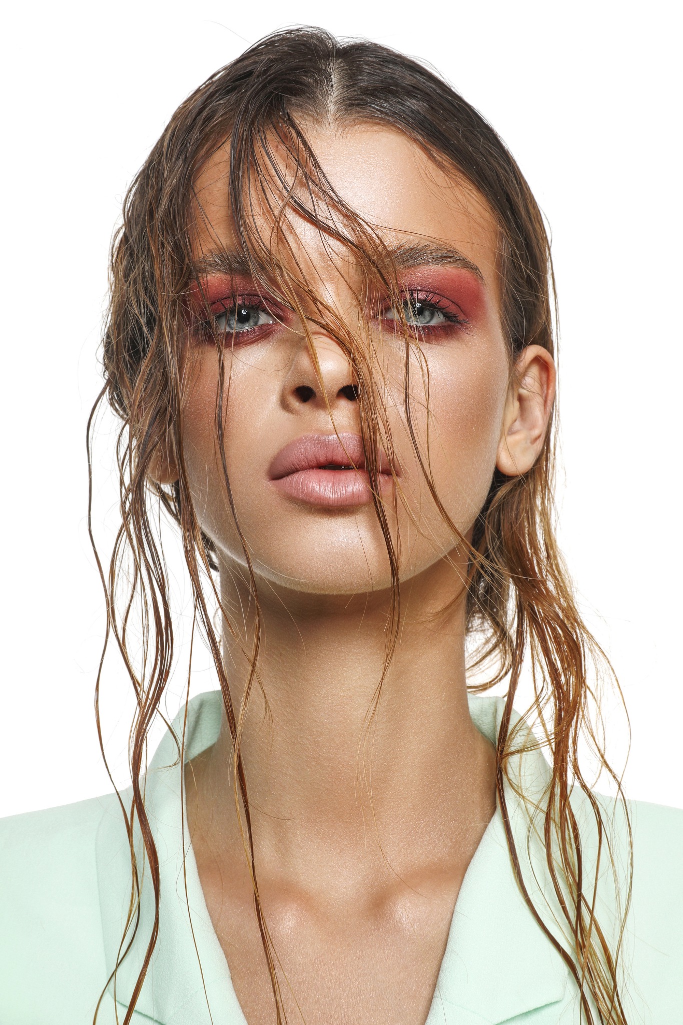 Portret kobiety z mokrymi włosami, intensywnym makijażem oczu w odcieniach czerwieni i delikatnymi ustami, ubrana w pastelową marynarkę na białym tle.