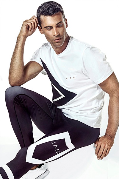 Mężczyzna w sportowym stroju: biała koszulka z czarnym wzorem i czarne legginsy z białymi elementami, siedzący na białym tle.