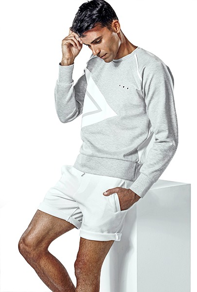 Mężczyzna w szarej bluzie z geometrycznym wzorem i białych szortach opiera się o biały sześcian, pozując w minimalistycznym studio.