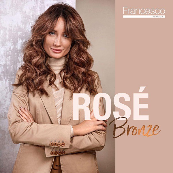 Portret kobiety z brązowymi, pofalowanymi włosami, ubrana w beżowy golf i marynarkę, z rękami skrzyżowanymi na piersiach, na tle z logo firmy i napisem 'ROSÉ Bronze'.