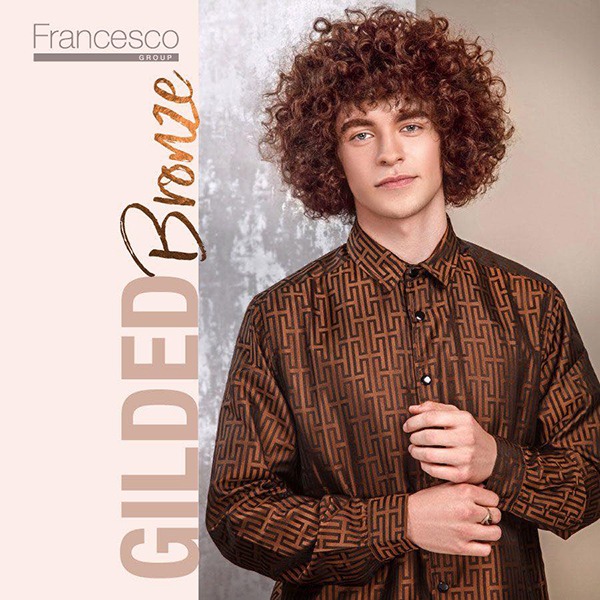 Mężczyzna z burzą rudych loków, ubrany w brązową koszulę w geometryczne wzory, pozuje na tle z teksturą, z widocznym logo firmy Francesco Group i napisem Gilded Bronze.