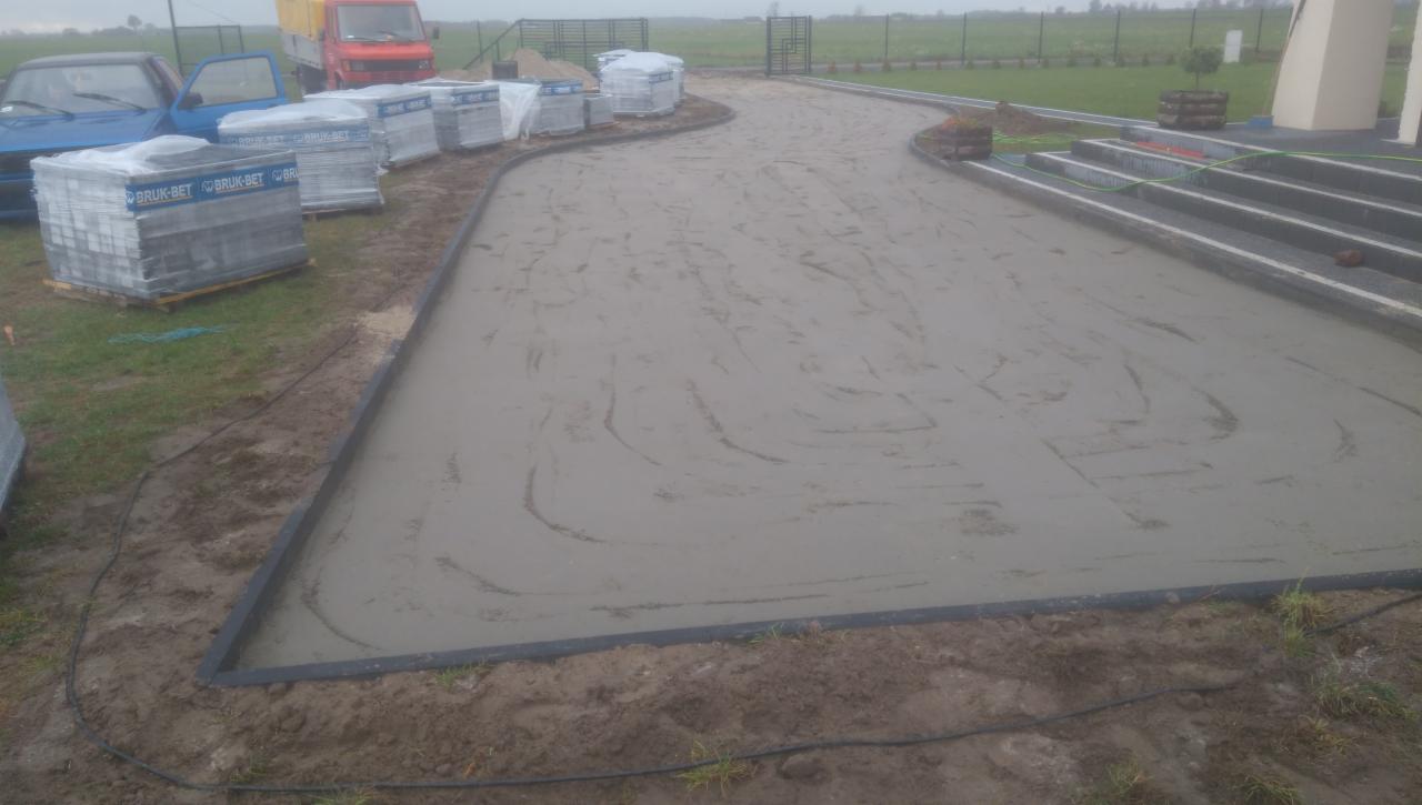 Przygotowanie podłoża pod nawierzchnię z kostki brukowej, widoczne obrzeża, przygotowany podkład cementowy i palety z kostką Bruk-Bet w tle.