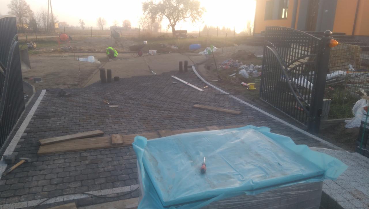 Układanie szarej kostki brukowej na podjeździe, widoczne narzędzia i materiały budowlane, brama wjazdowa z metaloplastyki w tle, pracownik w odblaskowej kamizelce i betoniarka w oddali, poranny...