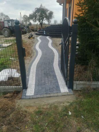Kaskadowa ścieżka z kostki brukowej w odcieniach szarości z białymi pasami prowadząca od bramy do schodów, w tle widoczny traktor i budynek mieszkalny.
