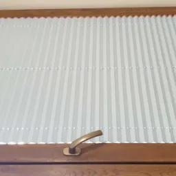 Białe plisowane przesłony okienne w drewnianej ramie, widoczne od wewnątrz pomieszczenia, z brązową klamką.