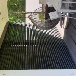 Zewnętrzne żaluzje fasadowe w kolorze antracytowym zamontowane na tarasie, z widocznym wiszącym fotelem i drewnianą podłogą tarasową