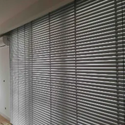 Szare poziome osłony okienne zamontowane na dużej powierzchni okiennej, widoczny klimatyzator i fragment parapetu pod oknem.