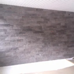 Ściana wewnętrzna wykończona szarą okładziną kamienną z nieregularnych, płaskich elementów, widok na fragment pokoju z białym sufitem panelowym i fragmentem okna po lewej stronie.