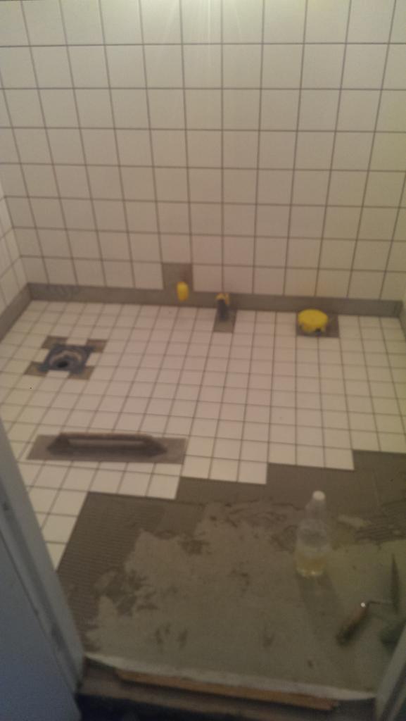 Remont łazienki w trakcie: układanie białych płytek kwadratowych na podłodze i ścianach kabiny prysznicowej, widoczny odpływ liniowy i punktowy, żółte gąbki, butelka z płynem i narzędzia glazurnicze.