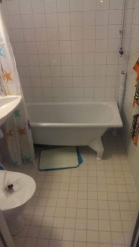 Wnętrze łazienki z białą wanną na nóżkach, toaletą i zasłoną prysznicową w gwiazdki, widok na białe kafelki ścienne i podłogowe, mały niebieski dywanik pod wanną.