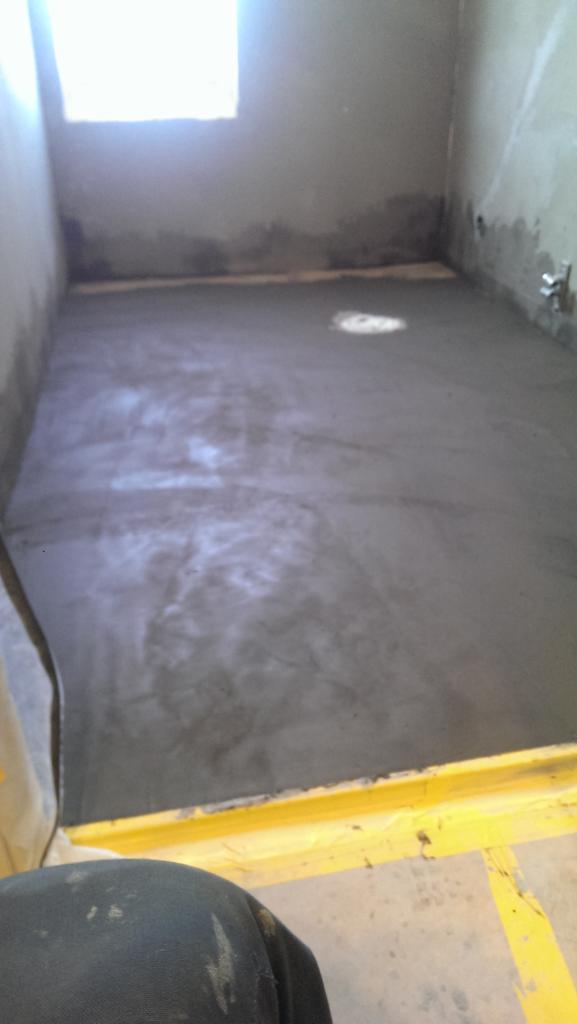 Wylewka betonowa w pomieszczeniu, widoczna instalacja w podłodze, ściany w stanie surowym, okno w tle, żółte elementy szalunku przy podłodze.
