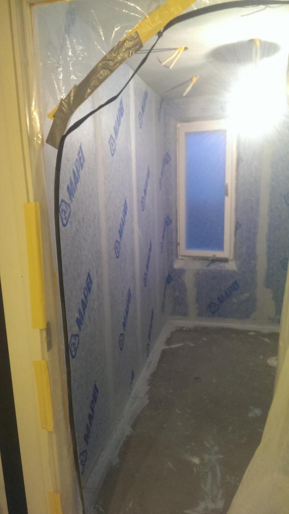 Przygotowanie poddasza do ocieplenia: ściany i okno zabezpieczone folią, widoczne logo MAPEI na materiale izolacyjnym, taśmy zabezpieczające w narożnikach.