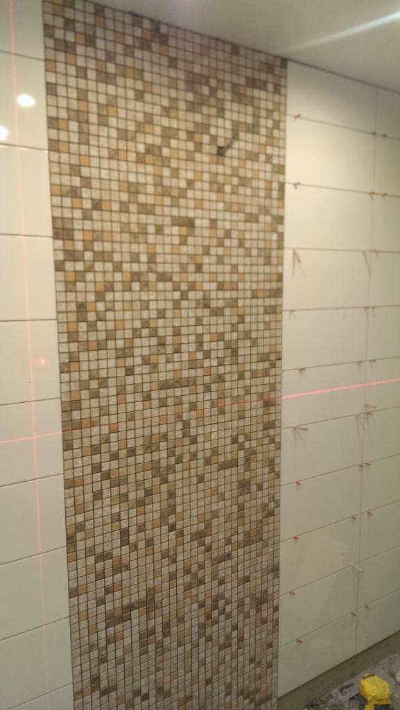 Układanie mozaiki w łazience z użyciem poziomicy laserowej, widoczne krzyżyki dystansowe między płytkami.