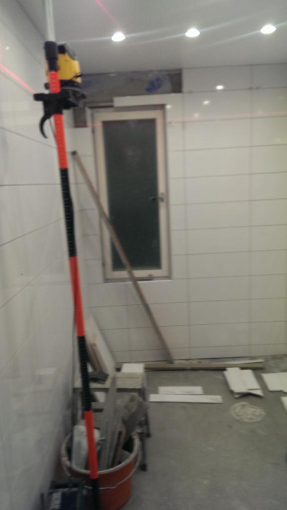 Wnętrze w trakcie remontu z białymi płytkami, widoczny laser krzyżowy na statywie, wiadro z narzędziami i materiałami budowlanymi, okno w tle.