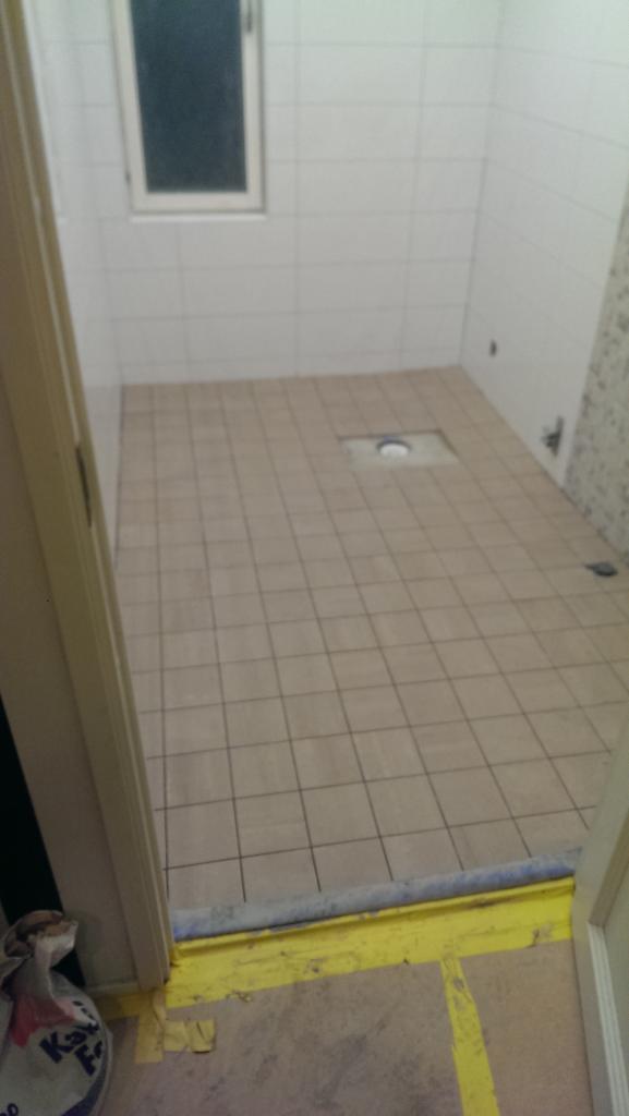 Wyłożona beżowymi płytkami podłoga w łazience z widocznym odpływem liniowym, zabezpieczona żółtą taśmą malarską przy progu drzwi.