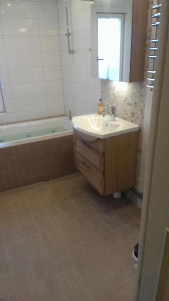 Nowoczesna łazienka z wanną obudowaną brązowymi płytkami, umywalką z szafką z jasnego drewna i lustrem z oświetleniem punktowym.