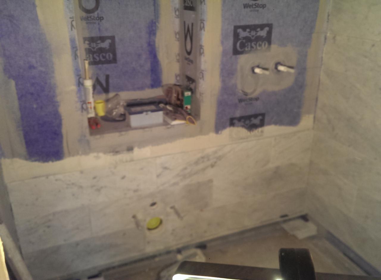 Wnętrze łazienki w trakcie remontu, widoczne białe płytki marmurowe na ścianach, zabezpieczone folią hydroizolacyjną z logo Casco i WetStop, przygotowane otwory na instalacje wodne i elektryczne...