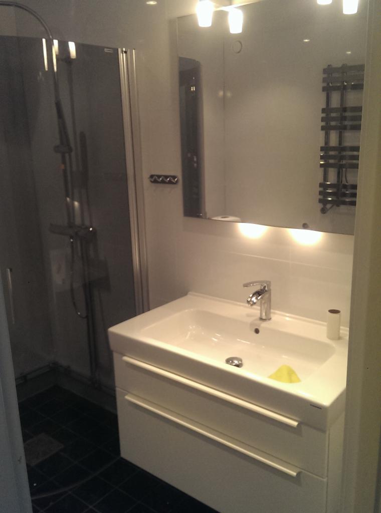 Nowoczesna łazienka z białą szafką pod umywalkę, lustrem z oświetleniem LED, kabiną prysznicową i ciemną podłogą z płytek.