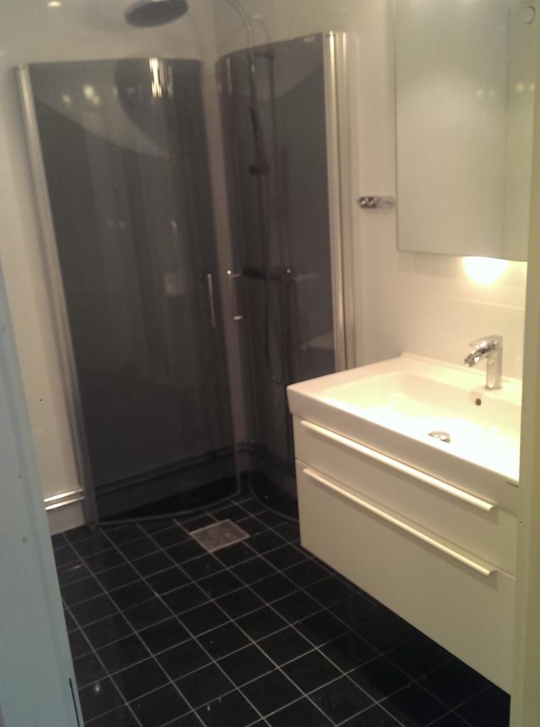 Nowoczesna łazienka z kabiną prysznicową z ciemnego szkła, białą szafką z umywalką i czarną podłogą z płytek.