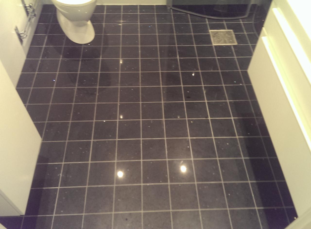 Ciemna, błyszcząca podłoga łazienki z kwadratowych płytek, widoczny fragment toalety i szafki umywalkowej.