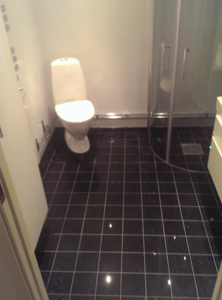 Wykończona łazienka z czarną, błyszczącą podłogą z płytek, białą toaletą i kabiną prysznicową z przezroczystego szkła.