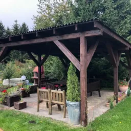 Zadaszona altana ogrodowa z ciemnego drewna, kryta blachą falistą, z meblami ogrodowymi wewnątrz, otoczona zielenią i kwiatami w donicach.