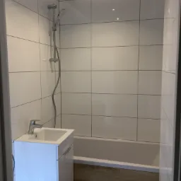 Wykończona łazienka z białymi, kwadratowymi płytkami na ścianach, wanną obudowaną panelami imitującymi drewno i białą szafką pod umywalką z chromowaną baterią oraz zestawem prysznicowym.