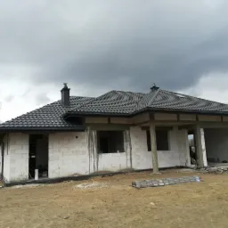 Dom w trakcie budowy z ciemnoszarą dachówką, widoczne rynny, komin i częściowo wykończone ściany z betonu komórkowego. Na pierwszym planie leży rozłożona drabina aluminiowa.