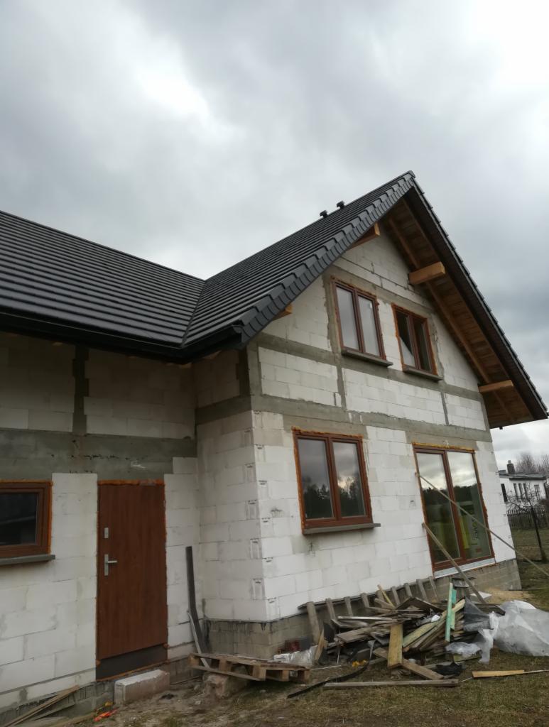 Dom w trakcie budowy z czarną dachówką, widoczne drewniane elementy konstrukcji dachu i niezakończone ściany z bloczków, pochmurne niebo w tle.