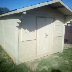 Surowy, drewniany domek narzędziowy z dwuspadowym dachem, umieszczony na trawniku, z widocznymi elementami konstrukcyjnymi i metalowym schowkiem w tle.