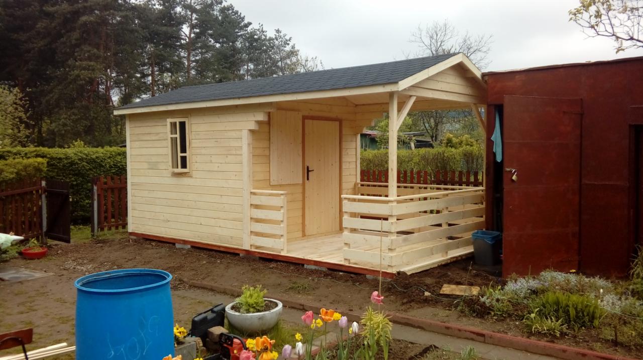 Surowy, drewniany domek narzędziowy z zadaszonym gankiem, jeszcze niepomalowany, z czarnym dachem, stojący na gruncie obok czerwonego garażu, w otoczeniu zieleni i kwiatów.