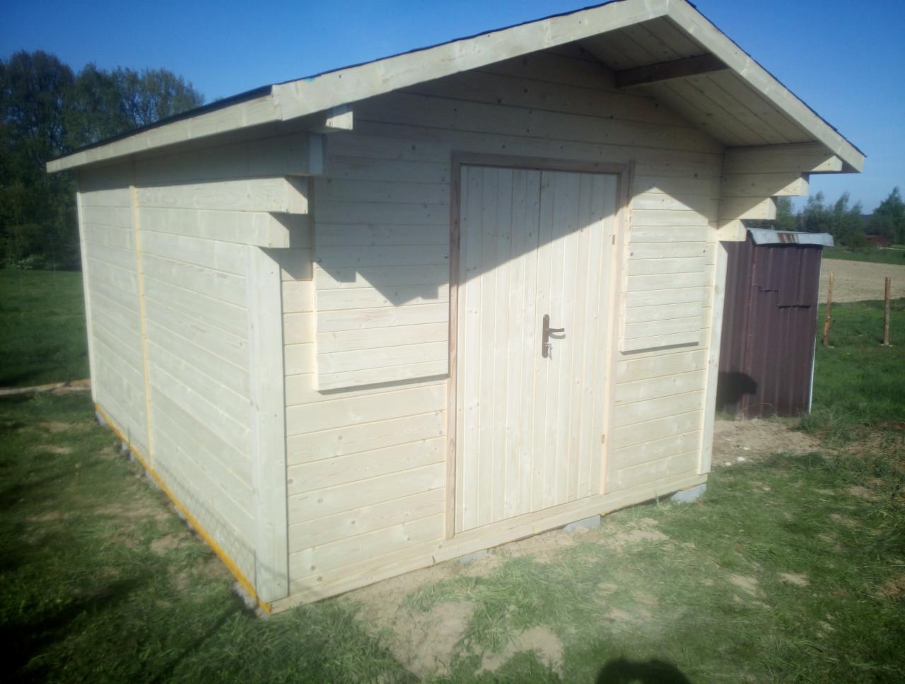 Surowy, drewniany domek narzędziowy z dwuspadowym dachem, umieszczony na trawniku, z widocznymi elementami konstrukcyjnymi i metalowym schowkiem w tle.