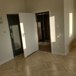 Wnętrze mieszkania po remoncie: widok na odnowioną podłogę w jodełkę, świeżo pomalowane ściany w odcieniach bieli, otwarte białe drzwi i kabina prysznicowa w tle oraz czarne drzwi wejściowe...