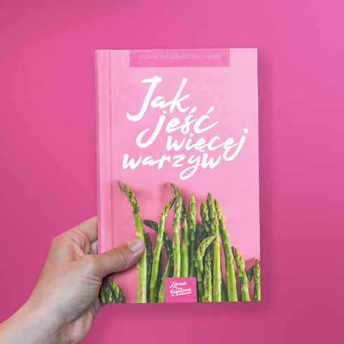 Ręka trzyma książkę o jedzeniu większej ilości warzyw, z okładką w odcieniach różu i ilustracją szparagów.