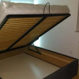 Uchylne łóżko tapicerowane z pojemnikiem, materac w folii ochronnej, widoczne listwy stelaża i mechanizm podnoszenia.