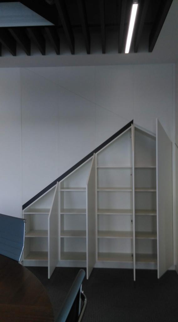Biała szafa wnękowa z półkami, dopasowana do skośnego sufitu, z czarnym obramowaniem górnej krawędzi, częściowo otwarta, widoczna z perspektywy pomieszczenia biurowego.